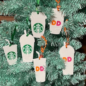 May include: Six white coffee cup ornaments with green and white Starbucks logos and orange and pink Dunkin' Donuts logos hanging on a green artificial Christmas tree.