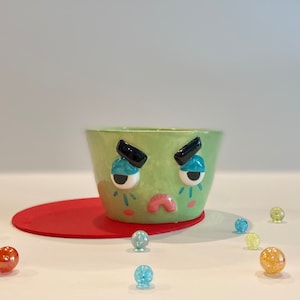 May include: A green ceramic bowl with a painted face. The face has black eyebrows, blue eyes, a pink nose, and a frown. The bowl is sitting on a red surface with colorful marbles scattered around it.