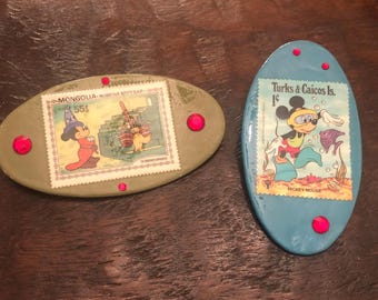 May include: Two oval-shaped ceramic magnets with images of Mickey Mouse. The first magnet is green and features Mickey Mouse dressed as a wizard with the text "MONGOLIA - монгол шуудан 55$". The second magnet is blue and features Mickey Mouse wearing a scuba mask and flippers with the text "Turks & Caicos Is. 1c MICKEY MOUSE".
