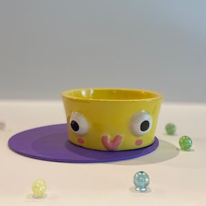 May include: A yellow ceramic bowl with a cartoon face, featuring two large eyes, a pink smile, and two small pink dots on the cheeks. The bowl is sitting on a purple surface.