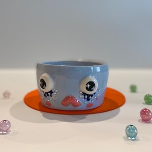 May include: A light blue ceramic bowl with a whimsical face. The bowl has large white eyes with black pupils, a pink nose, and a sparkly blue and white design around the eyes.