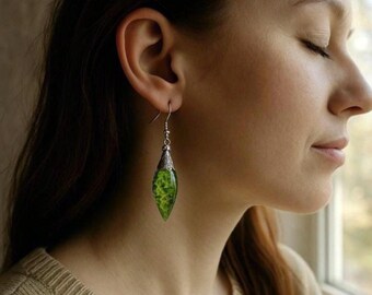 May include: A silver earring with a green, leaf-shaped pendant is worn by a woman. The pendant has a textured, marbled appearance with shades of green and yellow. The earring is suspended from the woman's ear.