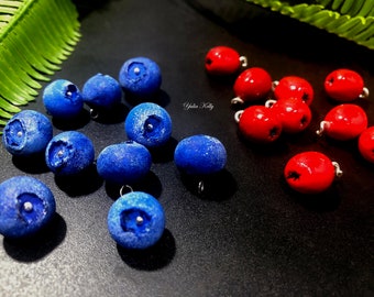 Può includere: Una collezione di ciondoli a forma di bacche blu e rosse con anelli d'argento. I ciondoli blu sono testurizzati e assomigliano ai mirtilli, mentre i ciondoli rossi sono lisci e assomigliano ai mirtilli rossi.