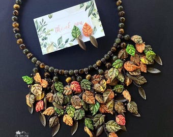 Può includere: Una collana di tendenza con una base in occhio di tigre e una frangia a cascata di foglie colorate. La collana è abbinata a orecchini coordinati con un design a foglia singola.