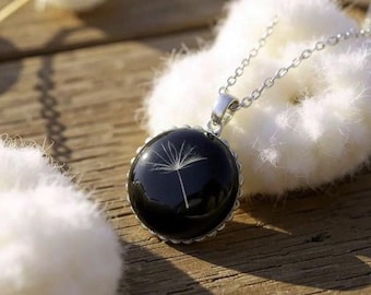 May include: A silver necklace with a round, black pendant. The pendant features a delicate white dandelion seed head encased in resin. The necklace is displayed on a wooden surface with white fluffy objects in the background.