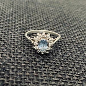 May include: A silver ring featuring a blue gemstone surrounded by smaller clear gemstones.