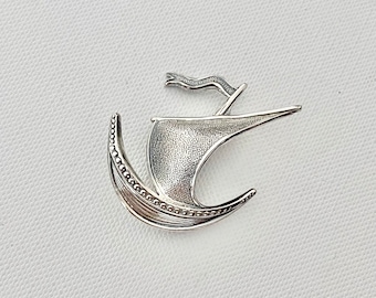 May include: A silver brooch in the shape of a stylized ship with a textured surface and a small sail.
