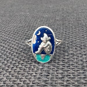 May include: Silver ring with an oval face depicting a seated figure under a crescent moon and stars. The background is dark blue, with a turquoise base. The ring has a detailed, whimsical design.