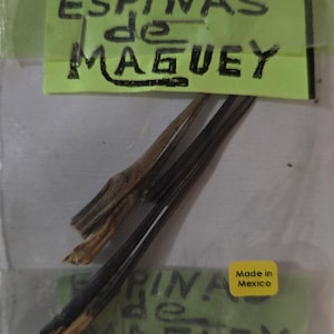 Op de afbeelding: Een doorzichtige plastic zak met verschillende donkerbruine en bruingele agavespines. De zak heeft een groen label met de tekst "ESPINAS de MAGUEY" in zwarte marker. Een kleine gele sticker leest "Made in Mexico".