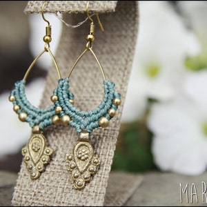May include: A pair of gold-toned earrings with teal macrame detailing and a decorative pendant. The earrings are hanging from a hessian display stand.