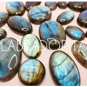 May include: A selection of polished labradorite stones in various oval shapes. The stones display iridescent blue, green, and gold hues, with a shimmering effect. The word "LABRADORITA" is written across the image in white capital letters.