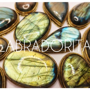 May include: An assortment of labradorite cabochons in gold-coloured settings. The stones exhibit iridescent colours of blue, green, and yellow. The word "LABRADORITA" is displayed across the image.