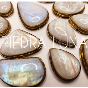 Op de afbeelding: Close-up van verschillende maansteen cabochons met een goudkleurige, touwachtige rand. De stenen zijn peer- en ovaalvormig, met een melkachtige witte basis en iriserende blauwe en gele flitsen. De tekst "PIEDRA LUNA" is over de afbeelding geplaatst.