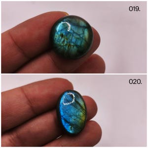 May include: Two polished labradorite gemstones. The top stone is round, displaying blue, green, and gold iridescence. The bottom stone is oval, with similar colors. The gemstones are held in a hand, with the numbers "019" and "020" visible.