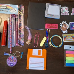 Op de afbeelding: Een verzameling briefpapier en accessoires in de stijl van de jaren 90, waaronder pennen, stickers, een sleutelhanger, een diskette en een notitieboek. De items zijn gerangschikt op een donker houten oppervlak, wat een nostalgische esthetiek oproept.
