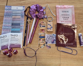 May include: A flat lay of various items including a purple hair scrunchie, a wooden bookmark with the text "Be Still And Know", a wooden keychain with a butterfly design, a pack of rose scented Epsom salts, a pack of tea bags, a purple lip balm, a pack of gold eye masks, a purple and silver pen, a black pen, a pink pen, a silver pen, a silver and black pen, a silver and black pencil, a purple bracelet, a silver paperclip, and a set of pink and grey sticky notes.
