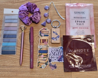 May include: A purple satin hair scrunchie, a set of pastel blue and pink sticky notes, two pens, a book stack sticker, a wooden bookmark with a dragonfly design, a key chain with a dragonfly design, a small bag of rose scented Epsom salt, and a small bag of gold eye masks.