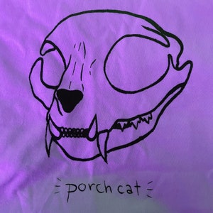 Könnte beinhalten: Weißes T-Shirt mit einer schwarzen Illustration eines Katzen-Schädels. Das Design enthält den Text "porch cat" in handschriftlicher Form unter dem Schädel. Das Bild ist mit einem violetten Farbton beleuchtet.