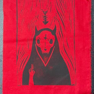 May include: Red fabric with a black graphic print. The print features a hooded cat skull with a cross on its forehead and a hand making a gesture. The background has a curtain design.