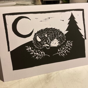 Pode incluir: Gravura em linóleo a preto e branco de uma raposa adormecida enrolada numa colina, com uma lua crescente e um pinheiro ao fundo. A raposa está rodeada por raminhos de bagas. A impressão está num cartão branco.