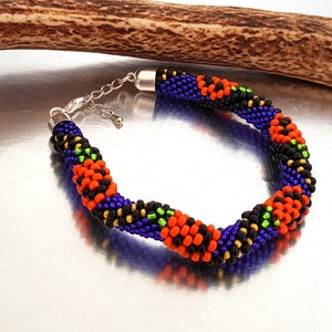 May include: A beaded bracelet with a black, orange, green, and purple color scheme. The bracelet is made with small seed beads and has a silver clasp.