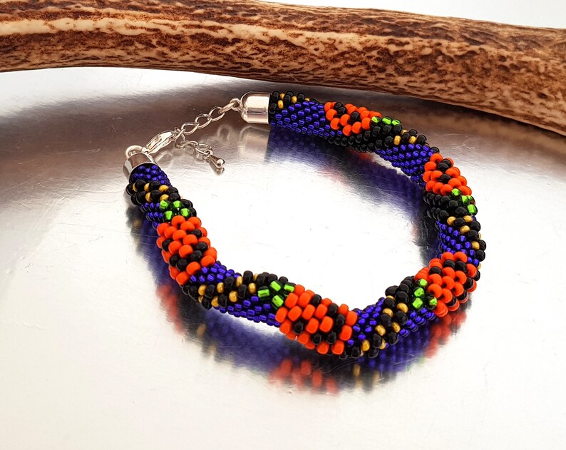 May include: A beaded bracelet with a black, orange, green, and purple color scheme. The bracelet is made with small seed beads and has a silver clasp.