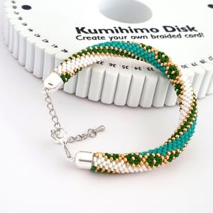 May include: A green, white, and gold beaded bracelet with a silver clasp. The bracelet is made using a Kumihimo disk, a tool for creating braided cords.