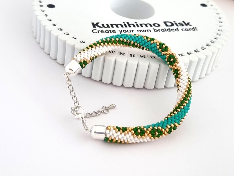 May include: A green, white, and gold beaded bracelet with a silver clasp. The bracelet is made using a Kumihimo disk, a tool for creating braided cords.