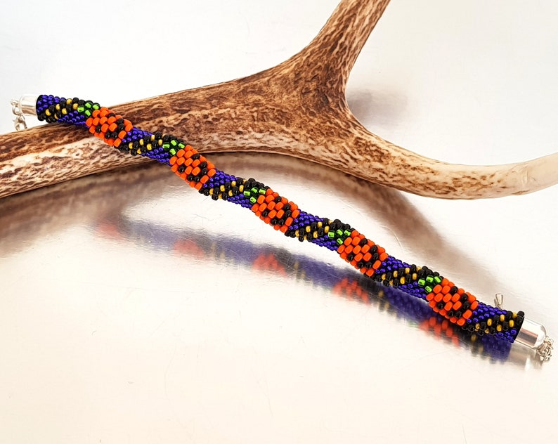 May include: A beaded bracelet with a repeating pattern of orange, black, green, and purple beads. The bracelet has silver clasps on each end.