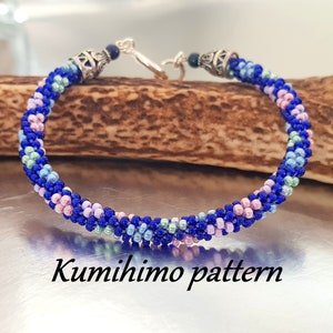 Könnte beinhalten: Ein blaues, rosa und grünes Perlenarmband mit silbernen Verschlüssen. Das Armband ist mit der Kumihimo-Technik hergestellt, einer traditionellen japanischen Flechtmethode. "Kumihimo pattern" steht unter dem Armband.