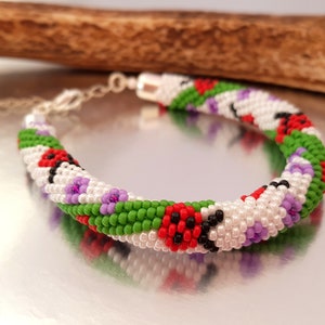 May include: A beaded bracelet featuring a repeating pattern of ladybugs, flowers, and green leaves. The bracelet is primarily white with red, black, green, and purple accents. The bracelet is approximately 7 inches in length.