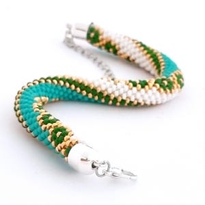 May include: A turquoise, green, and gold beaded bracelet with a silver clasp. The bracelet is made of small beads that are woven together in a spiral pattern.