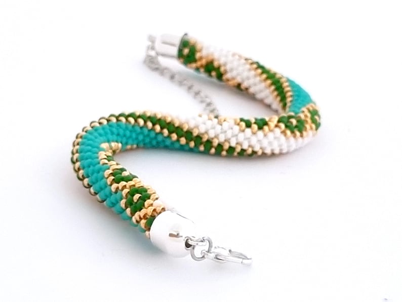 May include: A turquoise, green, and gold beaded bracelet with a silver clasp. The bracelet is made of small beads that are woven together in a spiral pattern.