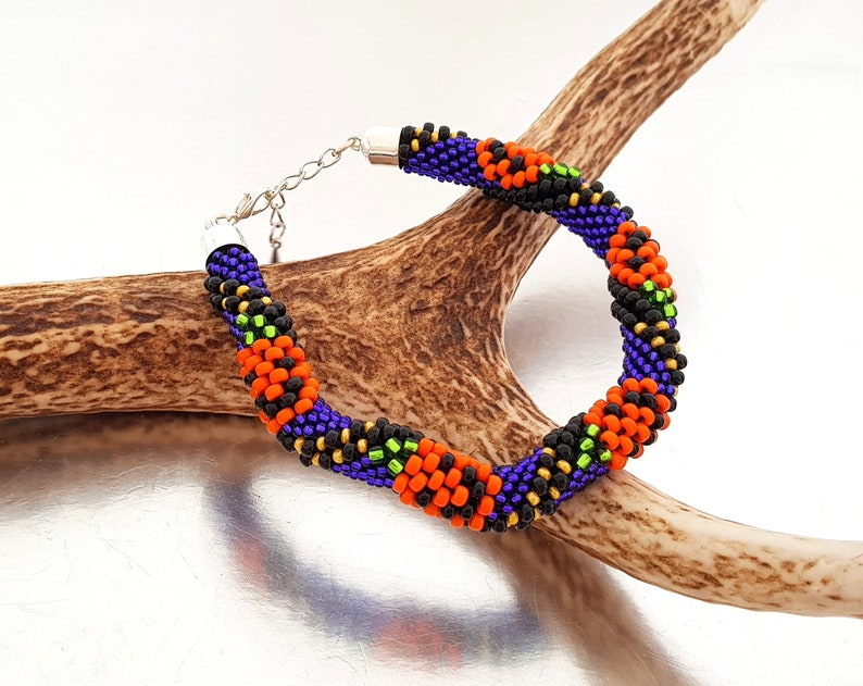 May include: A beaded bracelet with a repeating pattern of orange, black, green, and purple beads. The bracelet is on a brown antler.