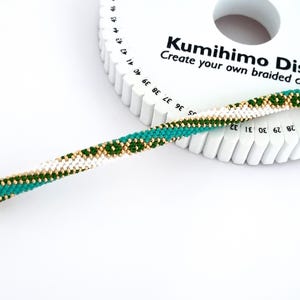 May include: A white, green, and gold braided cord made with a kumihimo disk. The disk is white with black numbers and the text "Kumihimo Disk Create your own braided cord!"