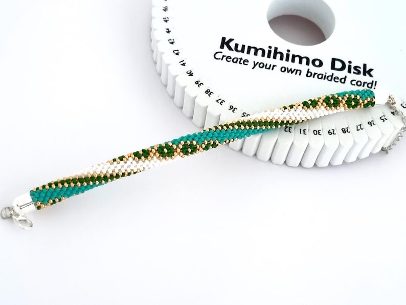 May include: A white, green, and gold braided cord made with a kumihimo disk. The disk is white with black numbers and the text "Kumihimo Disk Create your own braided cord!"