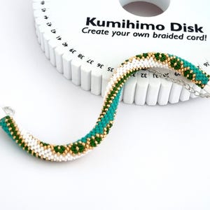 May include: A white Kumihimo Disk with green and gold braided cord. The disk has numbers around the edge and the text "Kumihimo Disk Create your own braided cord!" printed on it.