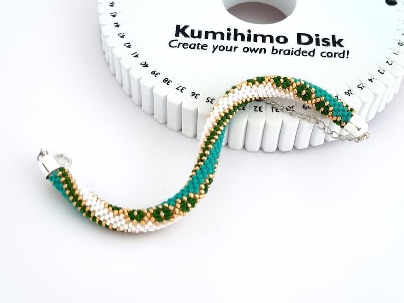 May include: A white Kumihimo Disk with green and gold braided cord. The disk has numbers around the edge and the text "Kumihimo Disk Create your own braided cord!" printed on it.