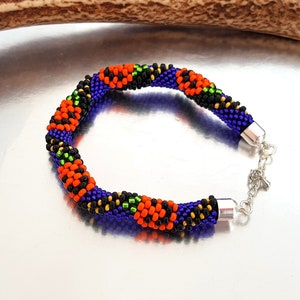 May include: A beaded bracelet with a black, orange, green, and purple color scheme. The bracelet has a silver clasp.