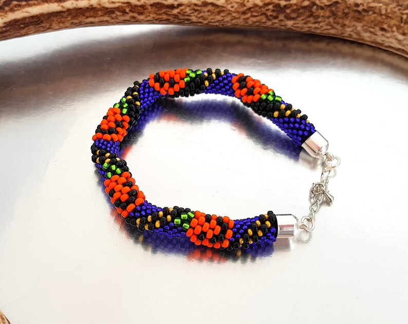 May include: A beaded bracelet with a black, orange, green, and purple color scheme. The bracelet has a silver clasp.