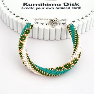 May include: A white, green, and gold braided bracelet made with a kumihimo disk. The disk is white with black numbers. The text "Kumihimo Disk" and "Create your own braided cord!" is printed on the disk.