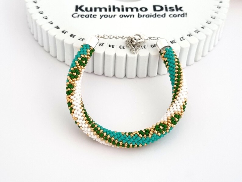 May include: A white, green, and gold braided bracelet made with a kumihimo disk. The disk is white with black numbers. The text "Kumihimo Disk" and "Create your own braided cord!" is printed on the disk.