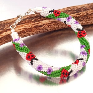 May include: A beaded bracelet with a colorful floral pattern. The bracelet features a white base with green, red, black, and purple accents. The bracelet has silver-colored end caps and a clasp. The bracelet is resting on a brown, textured surface.