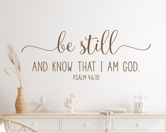 May include: A white wall with a brown vinyl decal that reads "be still and know that I am God. Psalm 46:10". The decal is above a white dresser with three drawers and a wicker chair with a white cushion.