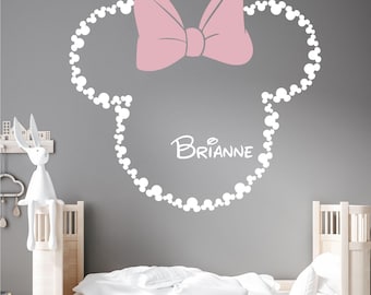 Könnte beinhalten: Weiße Vinyl-Wandaufkleber mit einer Mickey-Maus-Kopf-Silhouette mit einer rosa Schleife und dem Namen "BRIANNE" im Inneren. Der Aufkleber befindet sich an einer grauen Wand in einem Kinderzimmer.