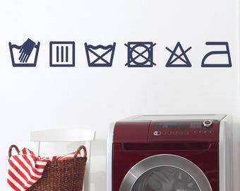 May include: A set of six laundry care symbols, including hand wash, machine wash, bleach, tumble dry, iron, and do not dry clean, are displayed on a white wall above a red and silver washing machine.