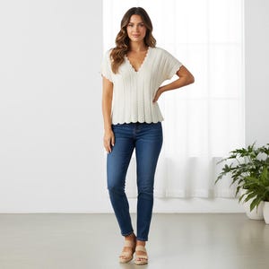 May include: A woman in a cream-coloured, short-sleeved sweater with a scalloped neckline and hem, and dark blue jeans. The sweater has a V-neck and a textured knit design. She is also wearing beige sandals.