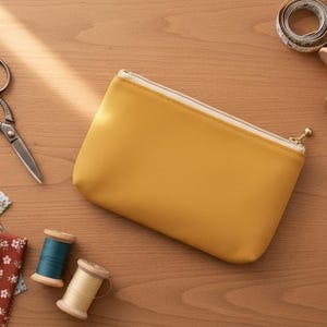 May include: Mustard yellow zippered pouch with a white zipper, surrounded by sewing supplies. Includes fabric scraps, scissors, spools of thread, a measuring tape, and pins, all on a wooden surface.