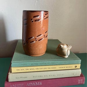 Puede incluir: Un jarrón cilíndrico de cerámica marrón con textura moteada y patrones de rayas horizontales de color marrón oscuro, acompañado de una pequeña figura de pájaro color canela, sobre una pila de cuatro libros titulados In the South of France, The Boy, the Mole, the Fox and the Horse, Before We Were Yours y Ansel Adams: An Autobiography.