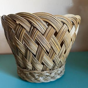 Op de afbeelding: Een ronde mand met open bovenkant, gemaakt van lichtbruin, natuurkleurig gevlochten riet. De mand heeft een brede, uitlopende rand en een dicht, ingewikkeld diagonaal vlechtpatroon over het hele lichaam, afgewerkt met een strakke, gevlochten rand.